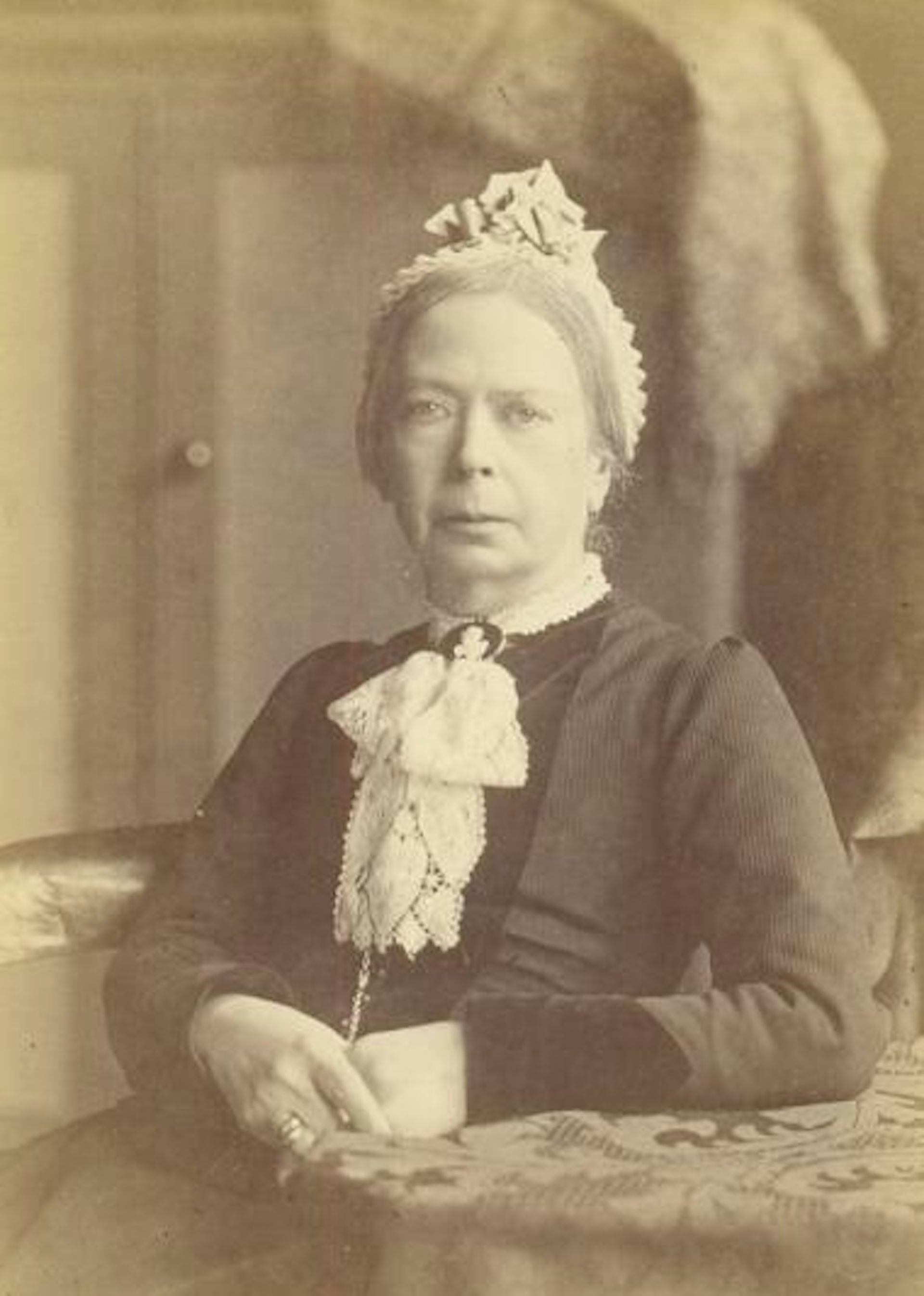 A sepia toned image of a serious-looking Victorian woman sitting with hands clasped.