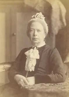 A sepia toned image of a serious-looking Victorian woman sitting with hands clasped.
