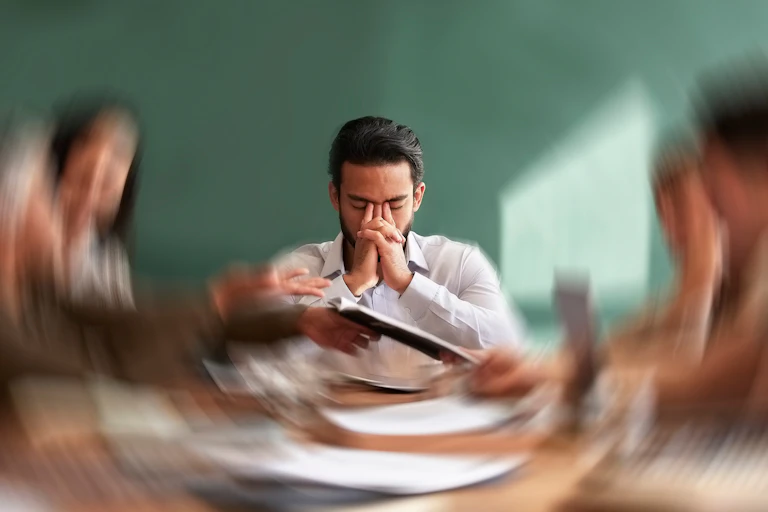 Homem estressado sentado à mesa, com pessoas em segundo plano, fora de foco.