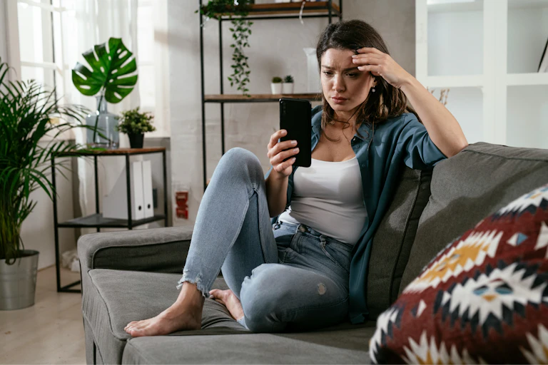 A woman sitting on a sofa looking at a phone screen with a puzzled expression