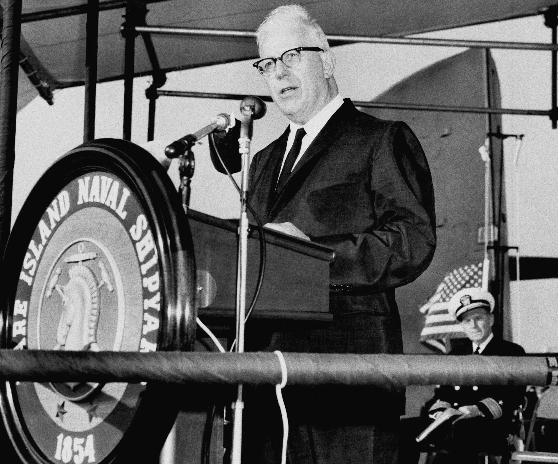 A man in a suit in tie in a black-and-white photo stands in front of a podium.