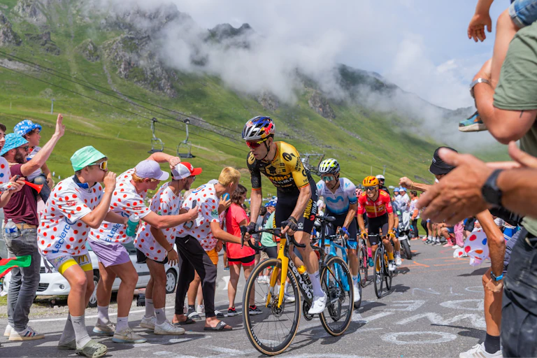 Unos ciclistas en una competición con montañas de fondo y aficionados a ambos lados de la carretera.