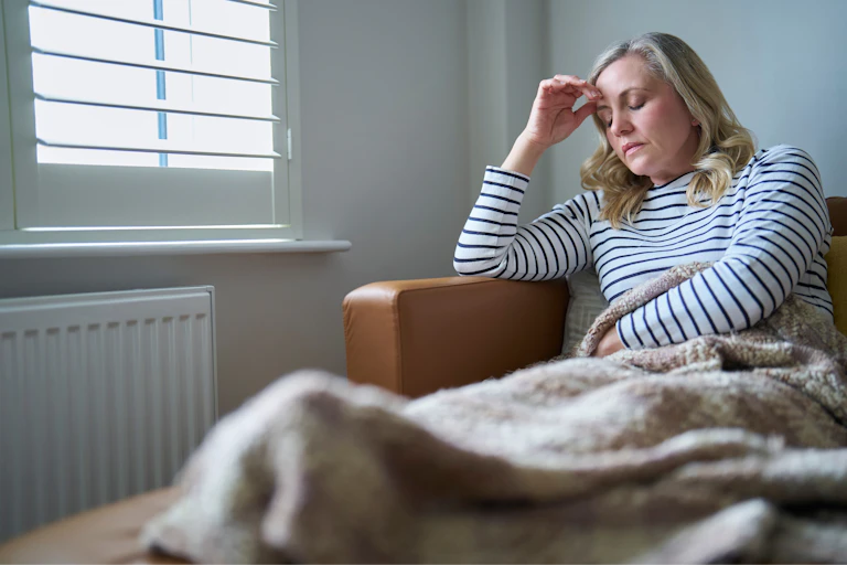 Ill woman lying on sofa, holds her head while closing her eyes. She looks exhausted.