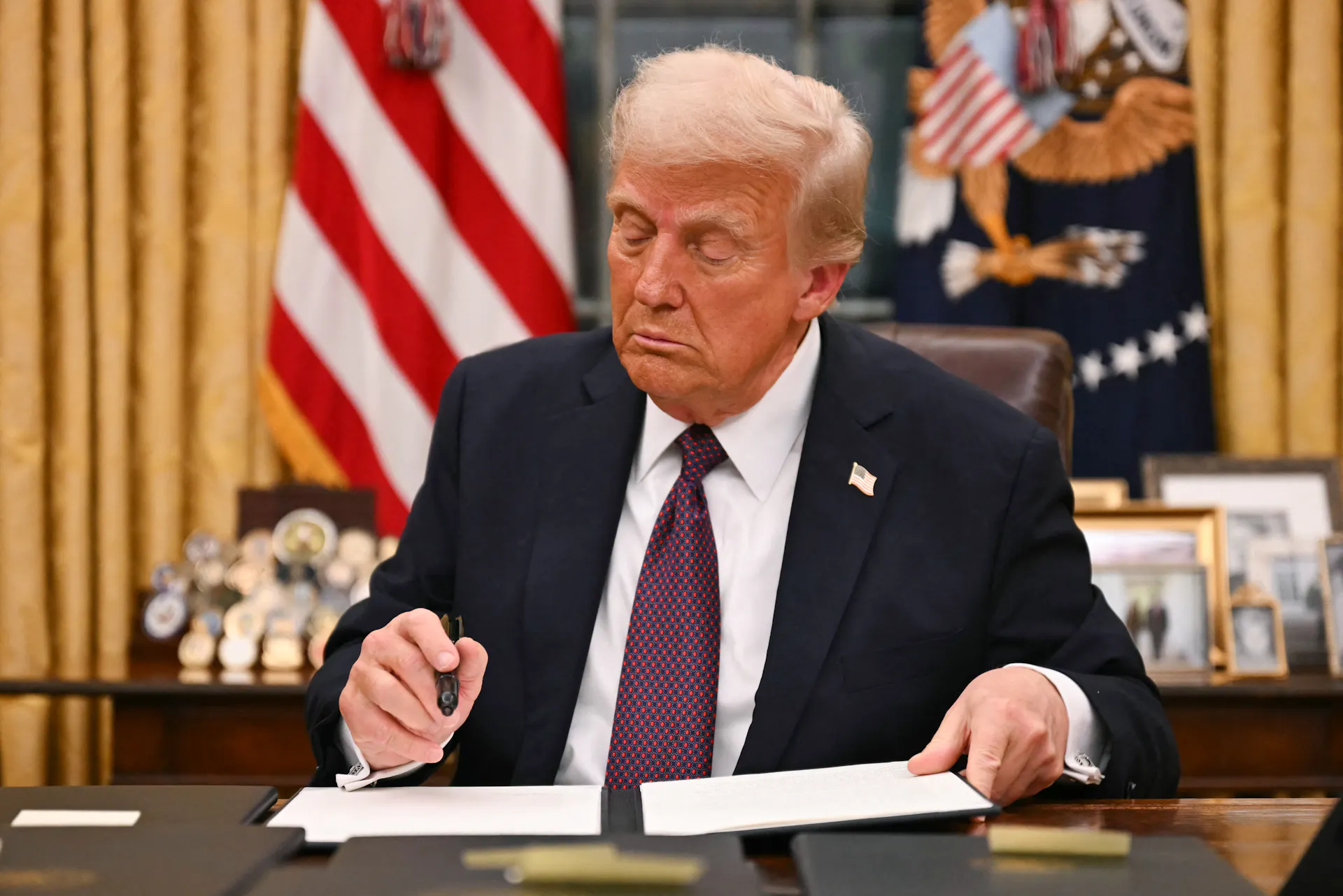 A man in a blue suit jacket sitting at a desk with a pen in his hand, looking at a folio.
