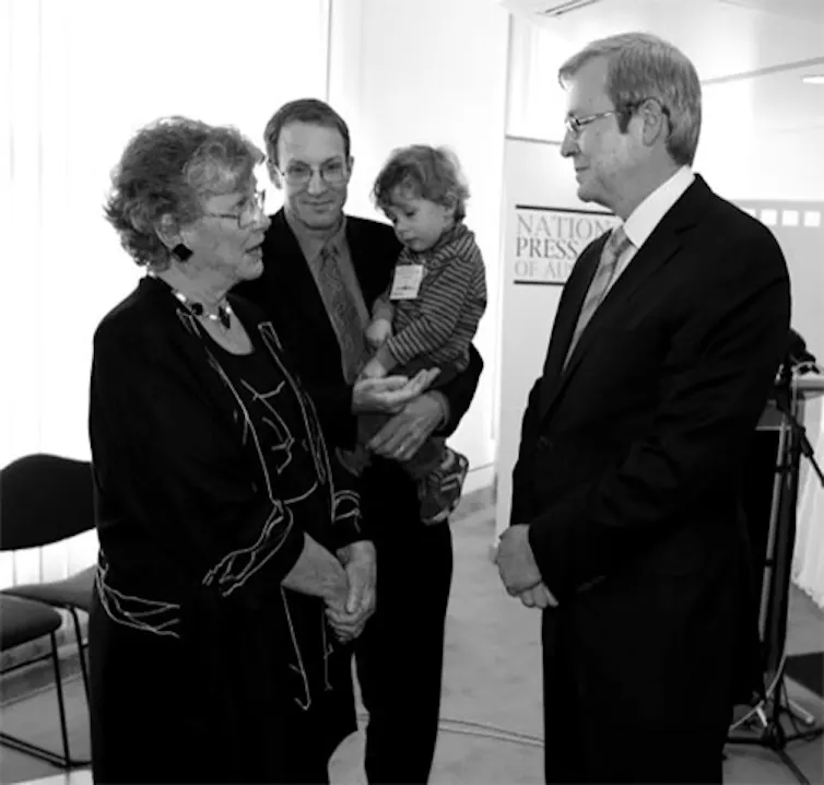 a woman talking to a man in a suit, with a man holding a baby in the background
