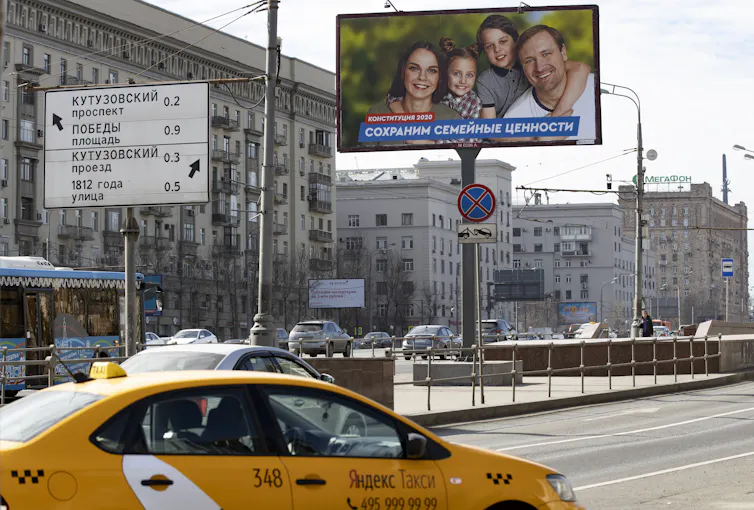 A yellow taxi drives past a billboard with a photo of a smiling woman, man and two children.