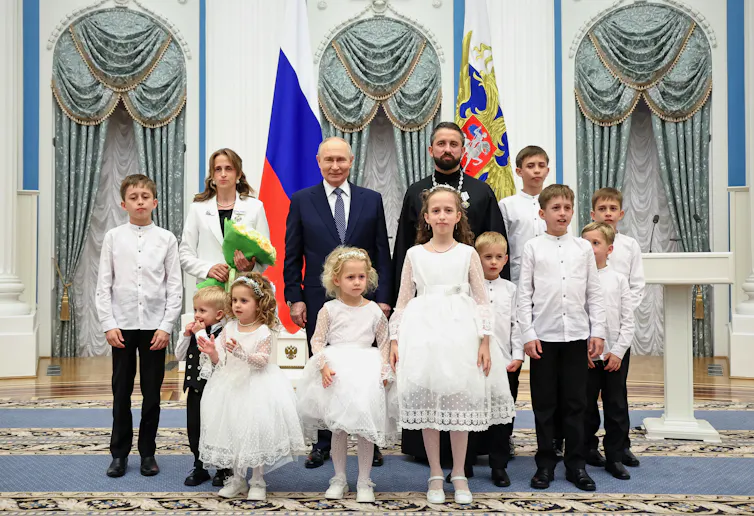Two men and a woman stand with 10 children in white outfits in an ornately decorated room.