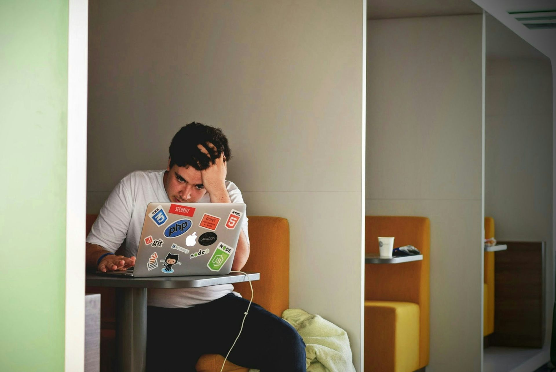 A young man in a white T-shirt stares into a stickered laptop at a coffee shop, head in hand.