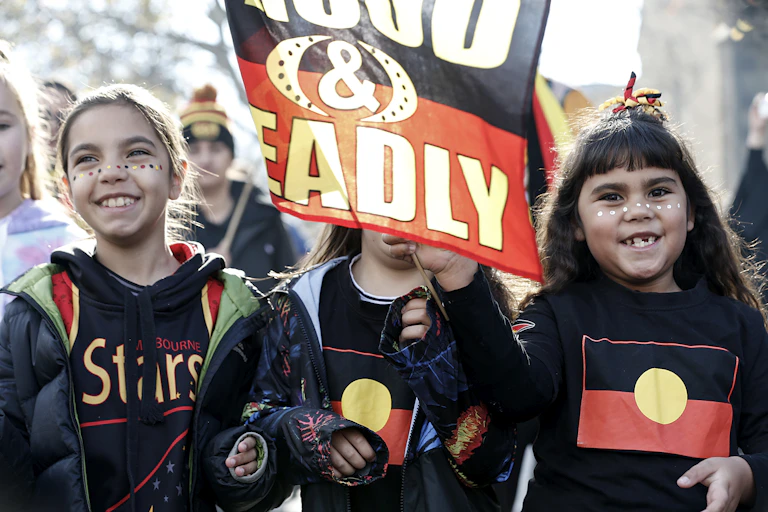Three young First Nations girls walk in a protest smiling and waving a flag that says proud and deadly