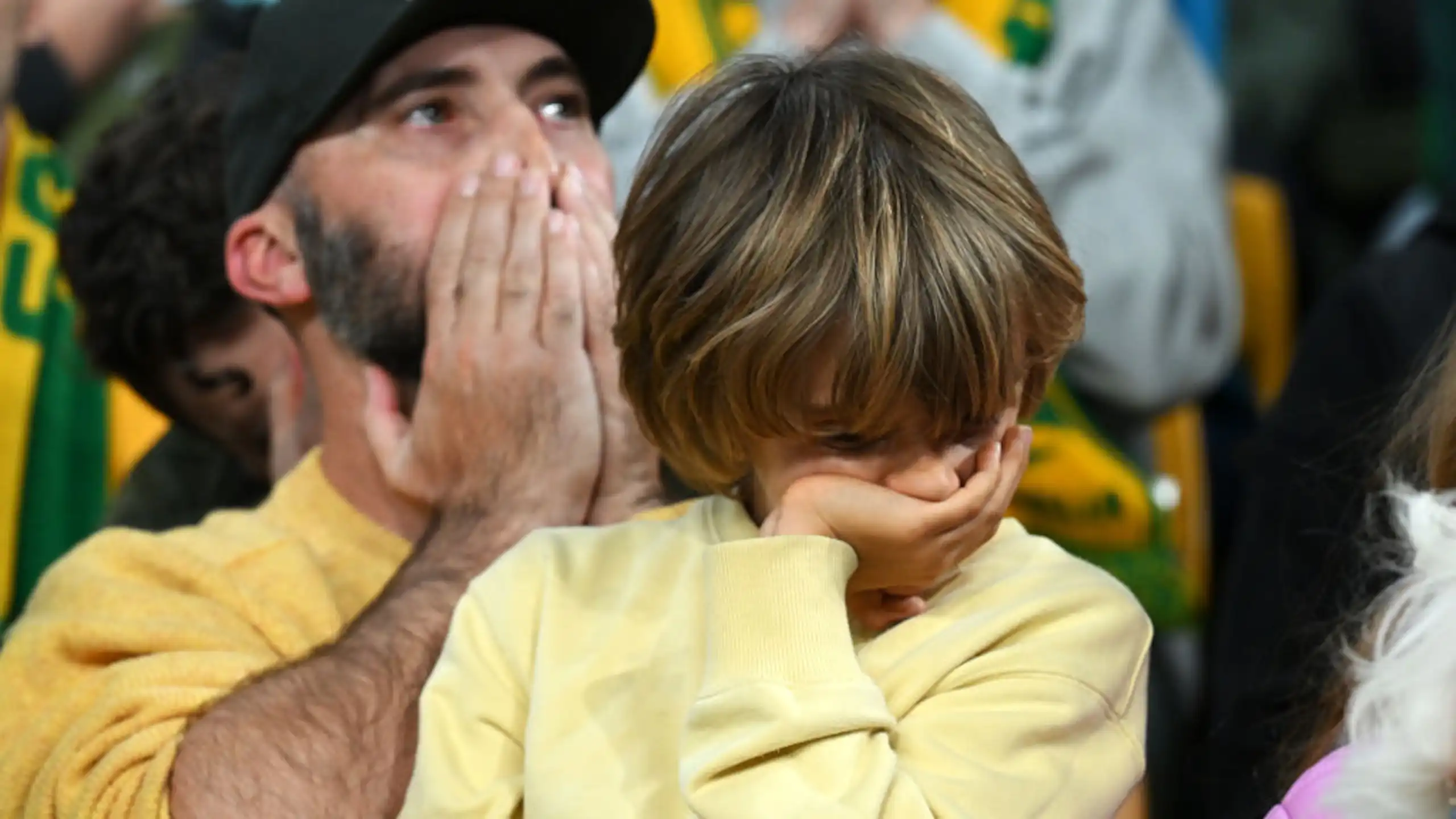 A young boy wearing a yellow jumper covers his mouth with his hand in disappointment while a man behind covers his own mouth in surprise.