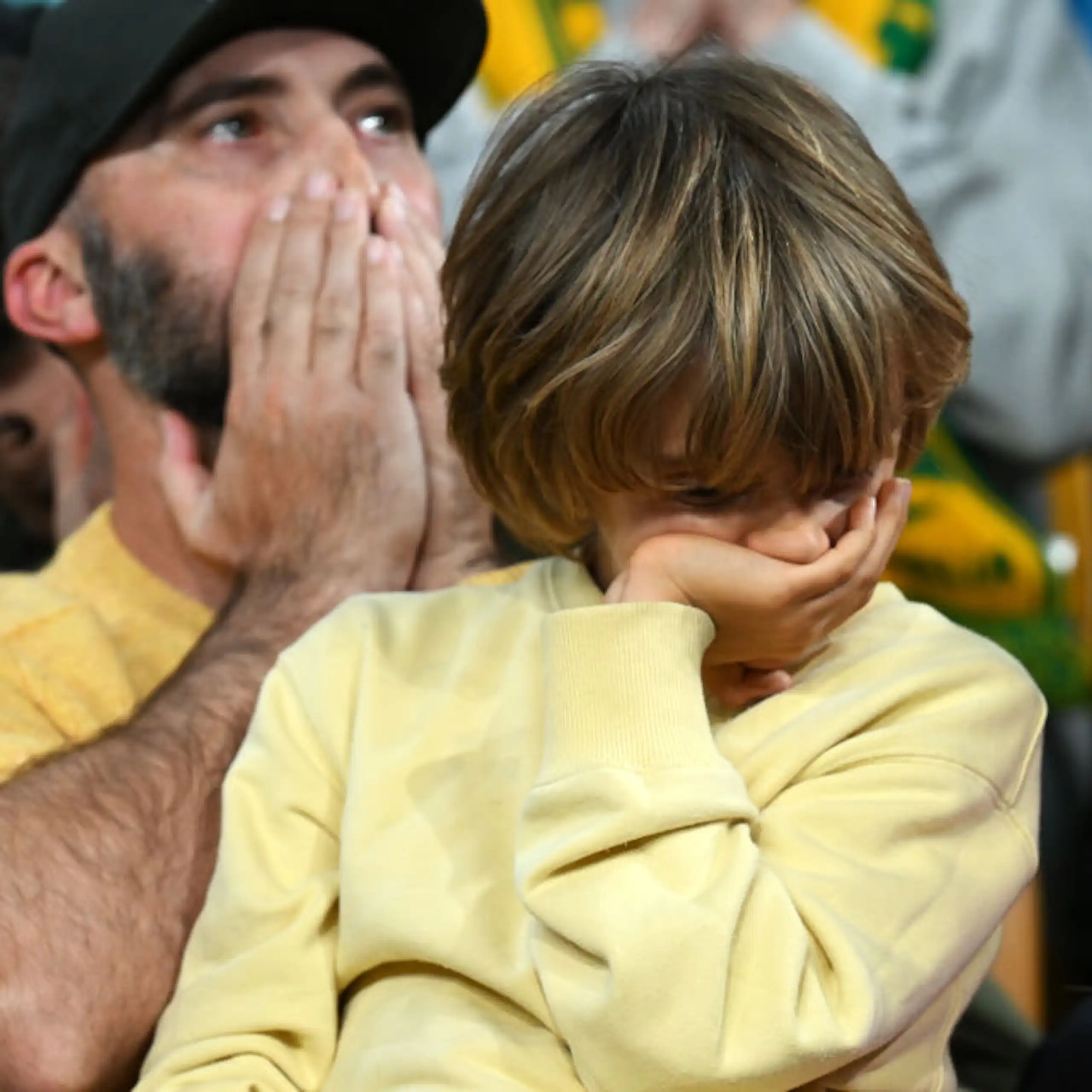 A young boy wearing a yellow jumper covers his mouth with his hand in disappointment while a man behind covers his own mouth in surprise.