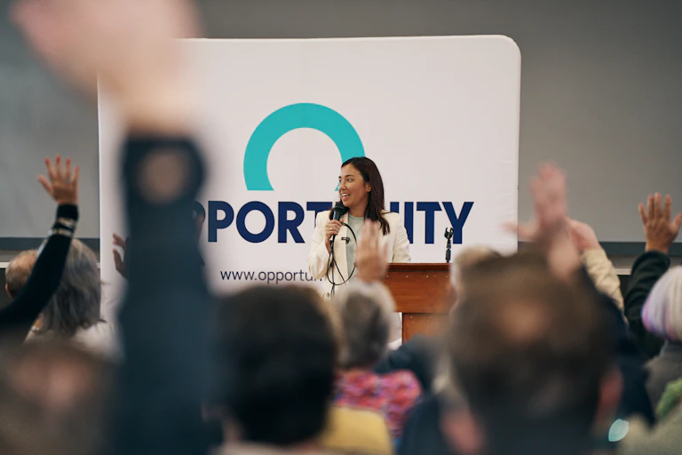 Woman speaks with a microphone in front of an Opportunity Party poster