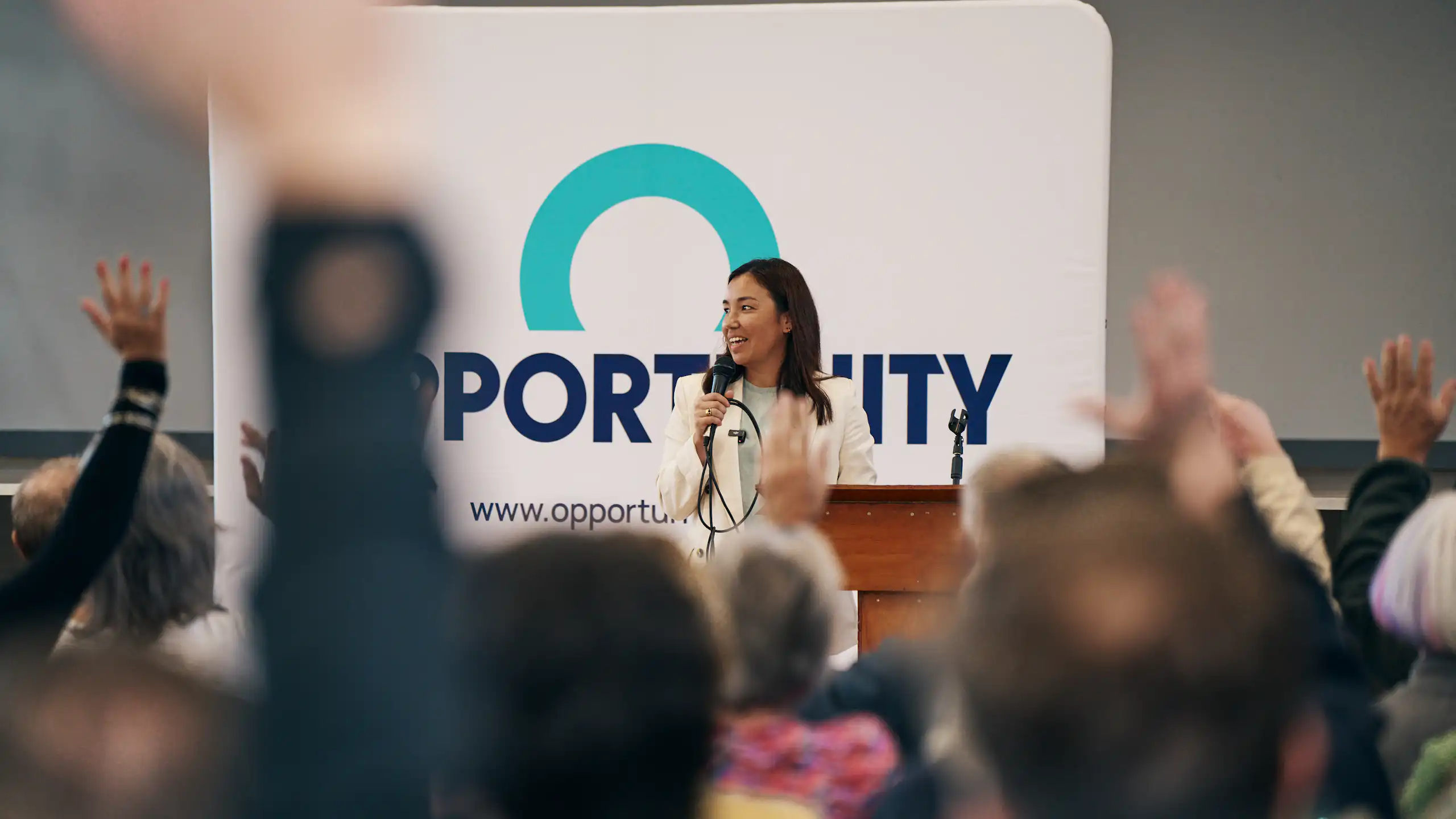 Woman speaks with a microphone in front of an Opportunity Party poster