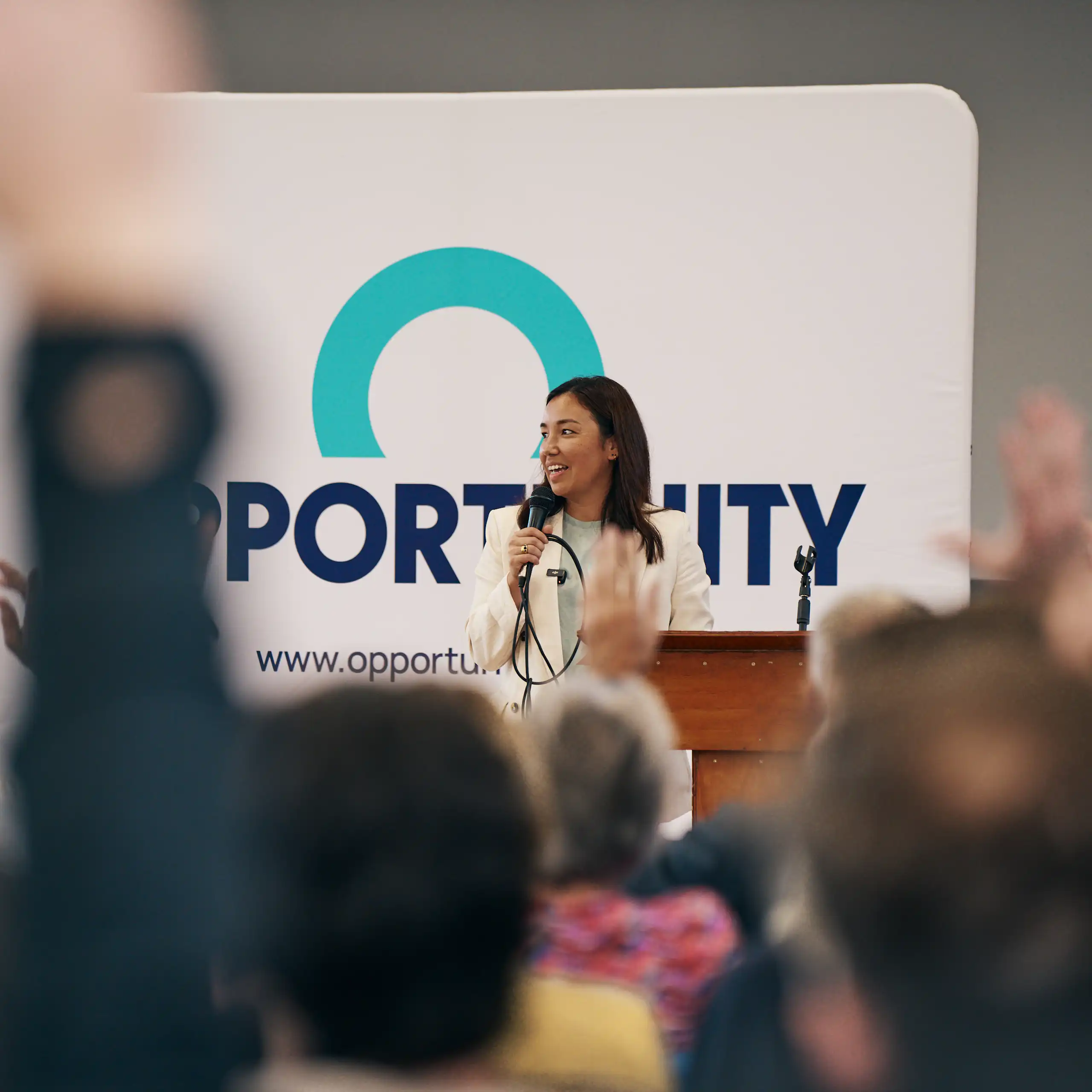 Woman speaks with a microphone in front of an Opportunity Party poster
