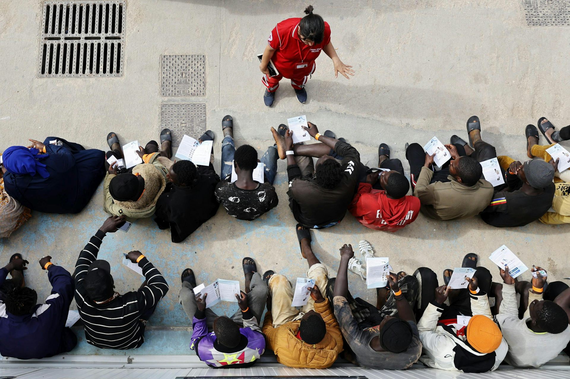 View from above of two rows of men sitting while an aid worker speaks to them