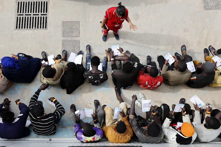 View from above of two rows of men sitting while an aid worker speaks to them