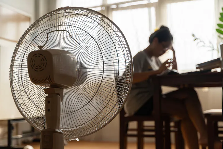 Woman sits at table at home with fan in foreground