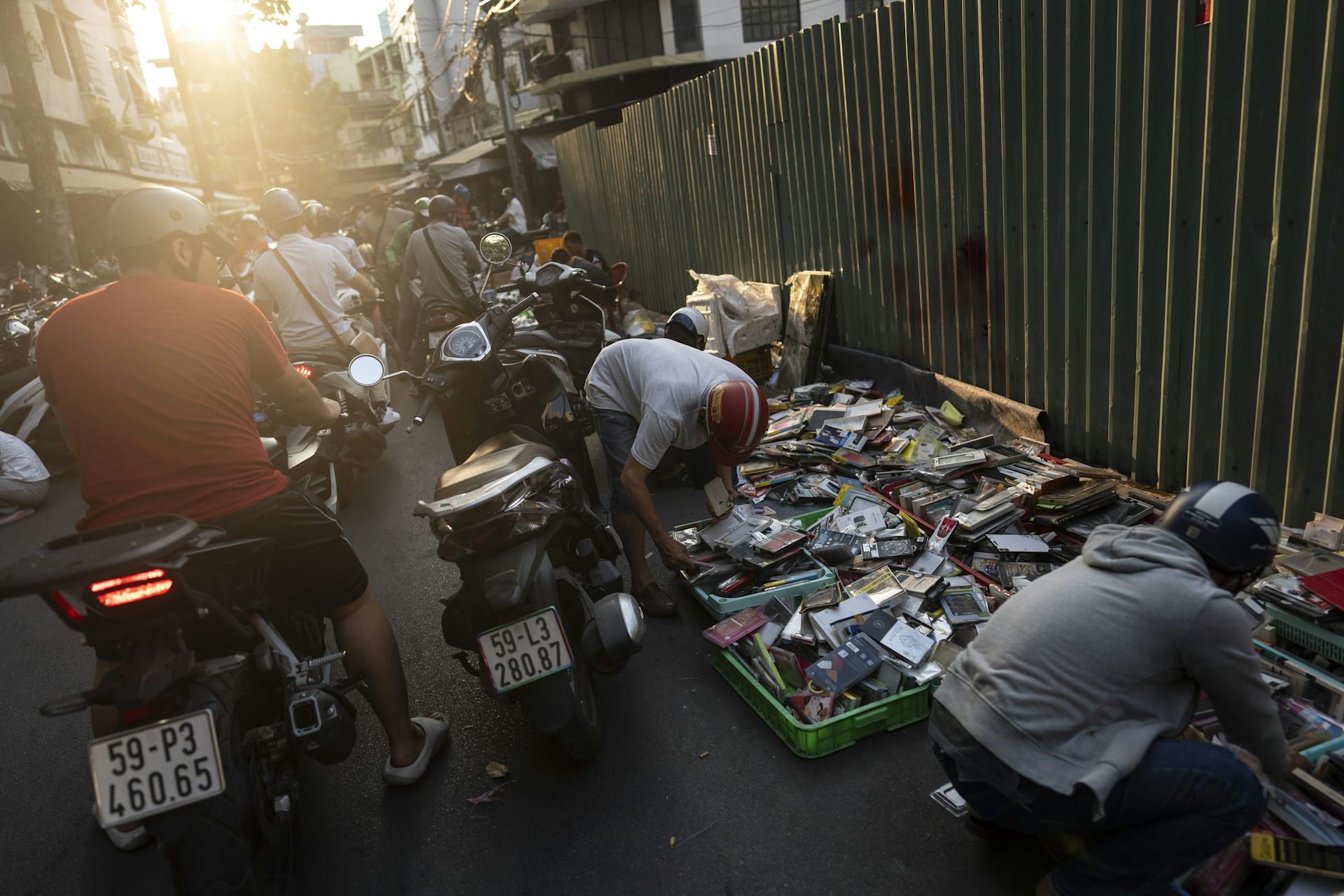 Vietnamese people in an alley rummage through boxes filled with electronic parts, while others ride by on motorcycles