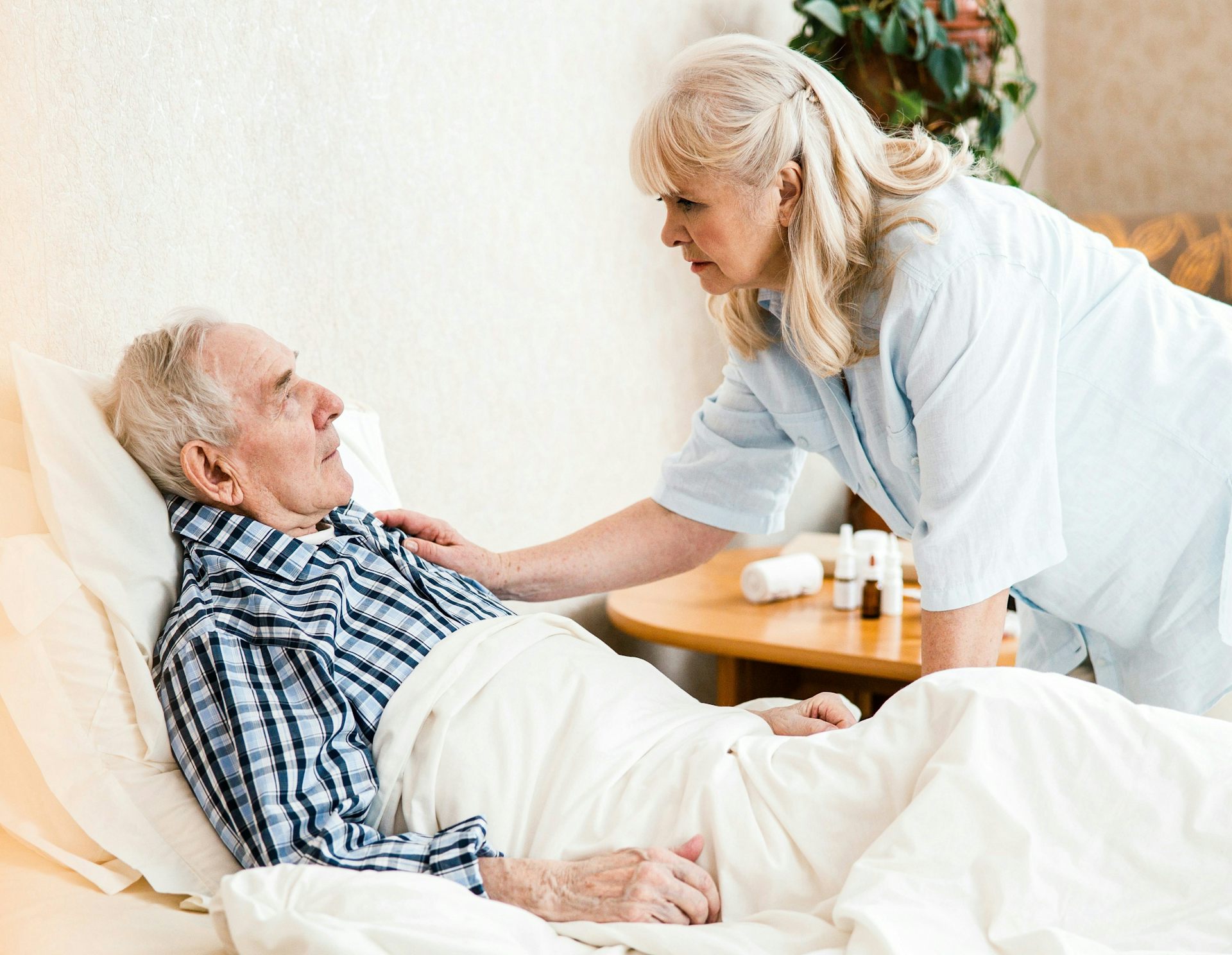 An elderly man in bed being cared for by a woman who is putting a hand on his shoulder.
