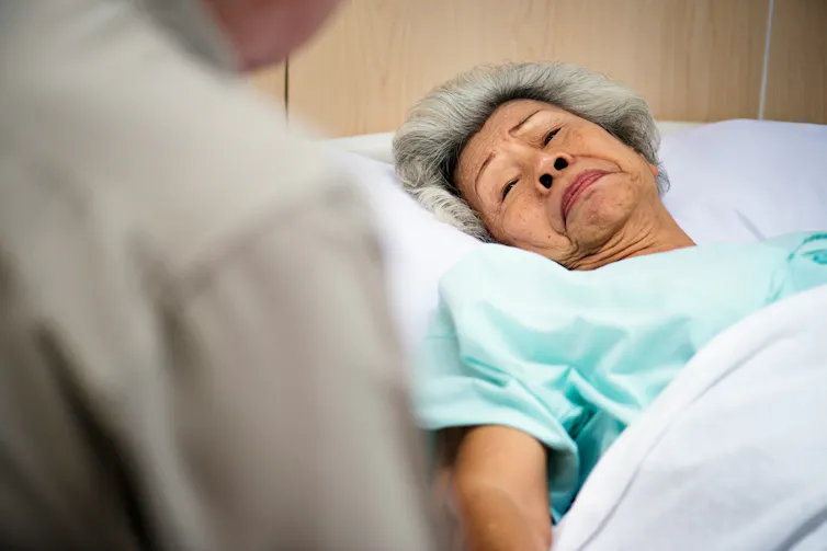 Primer plano de una anciana en la cama con una persona sentada al lado de su cama.
