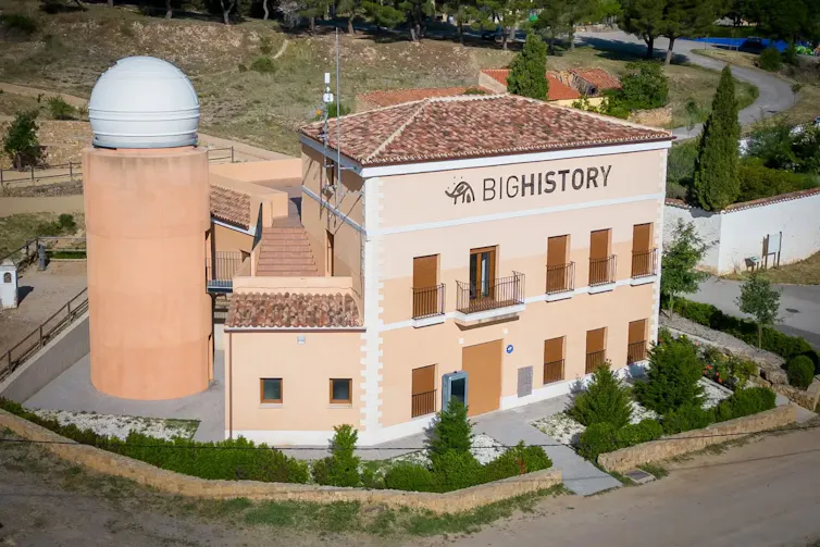 Un edificio de tres plantas con balcones en el que pone 'BIG HISTORY' rodeador por árboles y setos