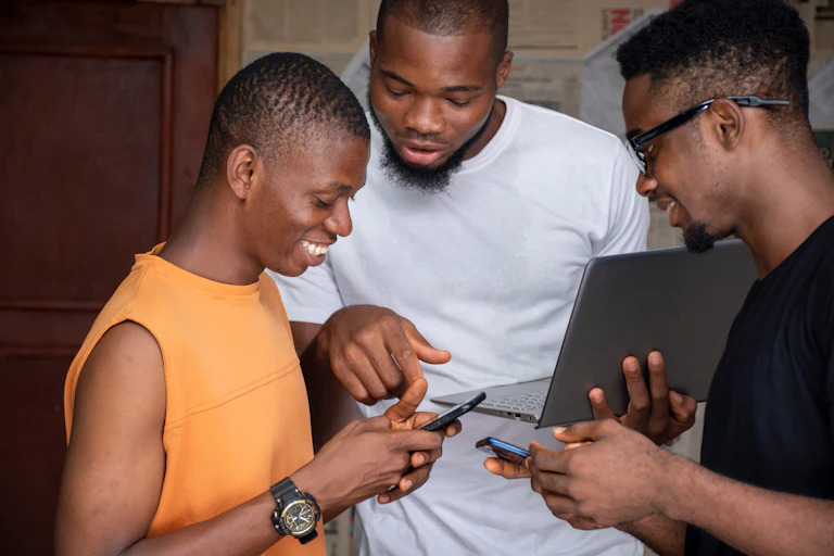 Three young Africans smile as they look at their mobile phones and laptop.
