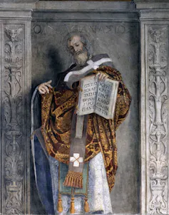 A bearded figure in an orange and brown robe gestures toward a large open book that he holds in one hand.