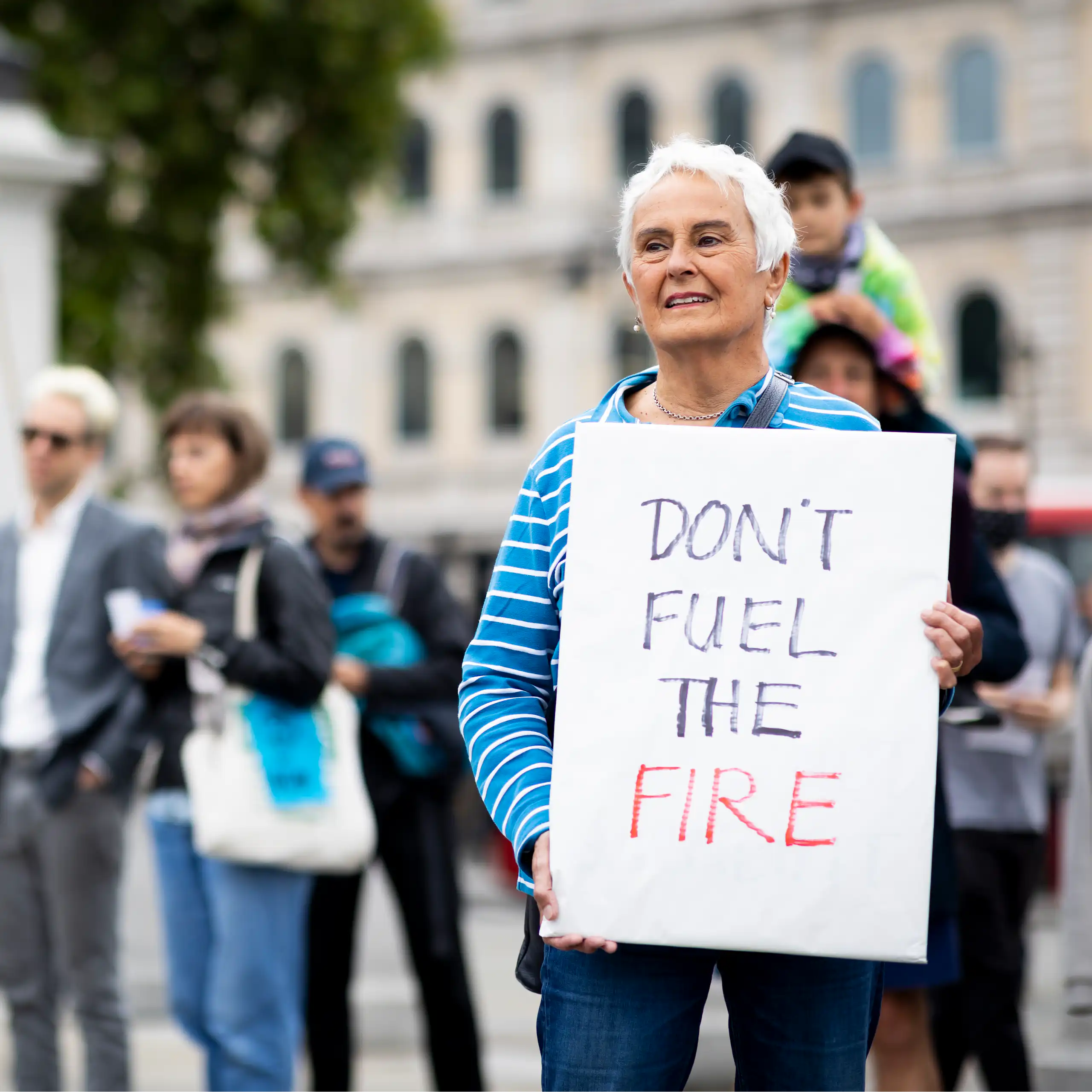 A woman with grey hair in a blue striped shirt protesting climate change holding a sign reading 'Don't fuel the fire'