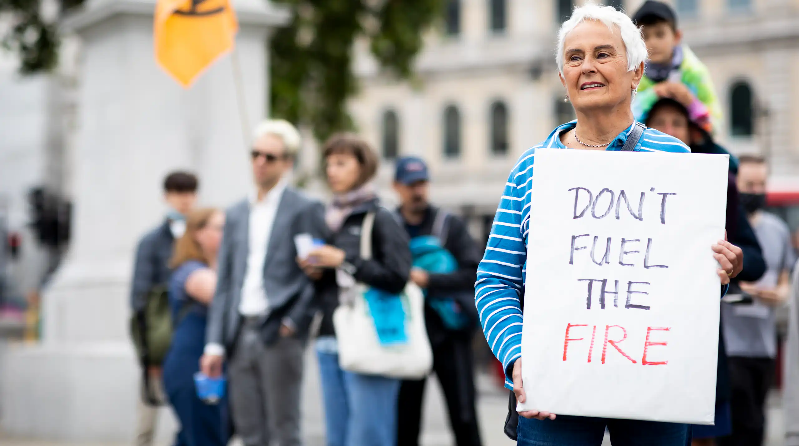 A woman with grey hair in a blue striped shirt protesting climate change holding a sign reading 'Don't fuel the fire'