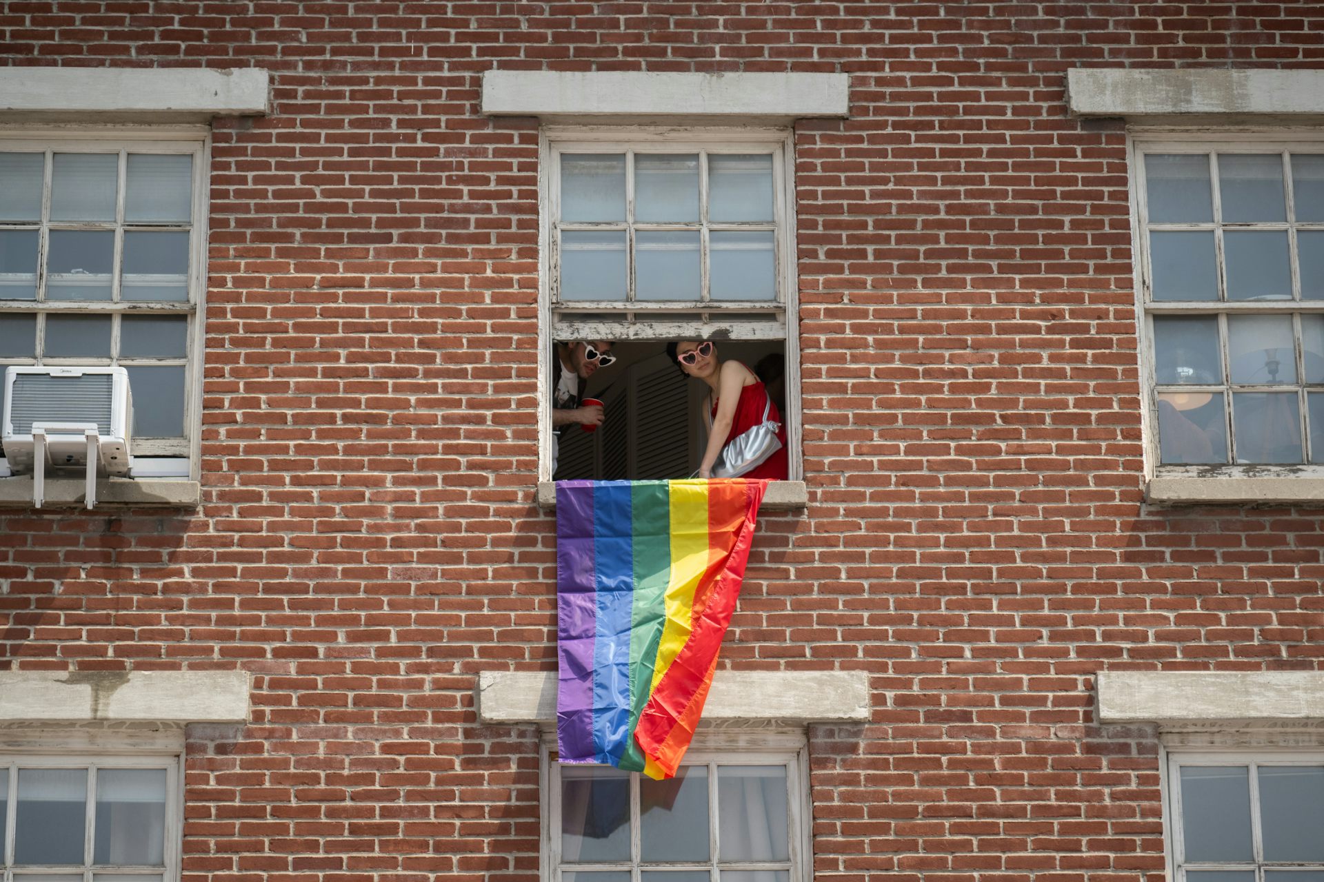 Two people wearing heart-shaped sunglasses lean out a window of a red brick building, a rainbow flag hanging from the sill.