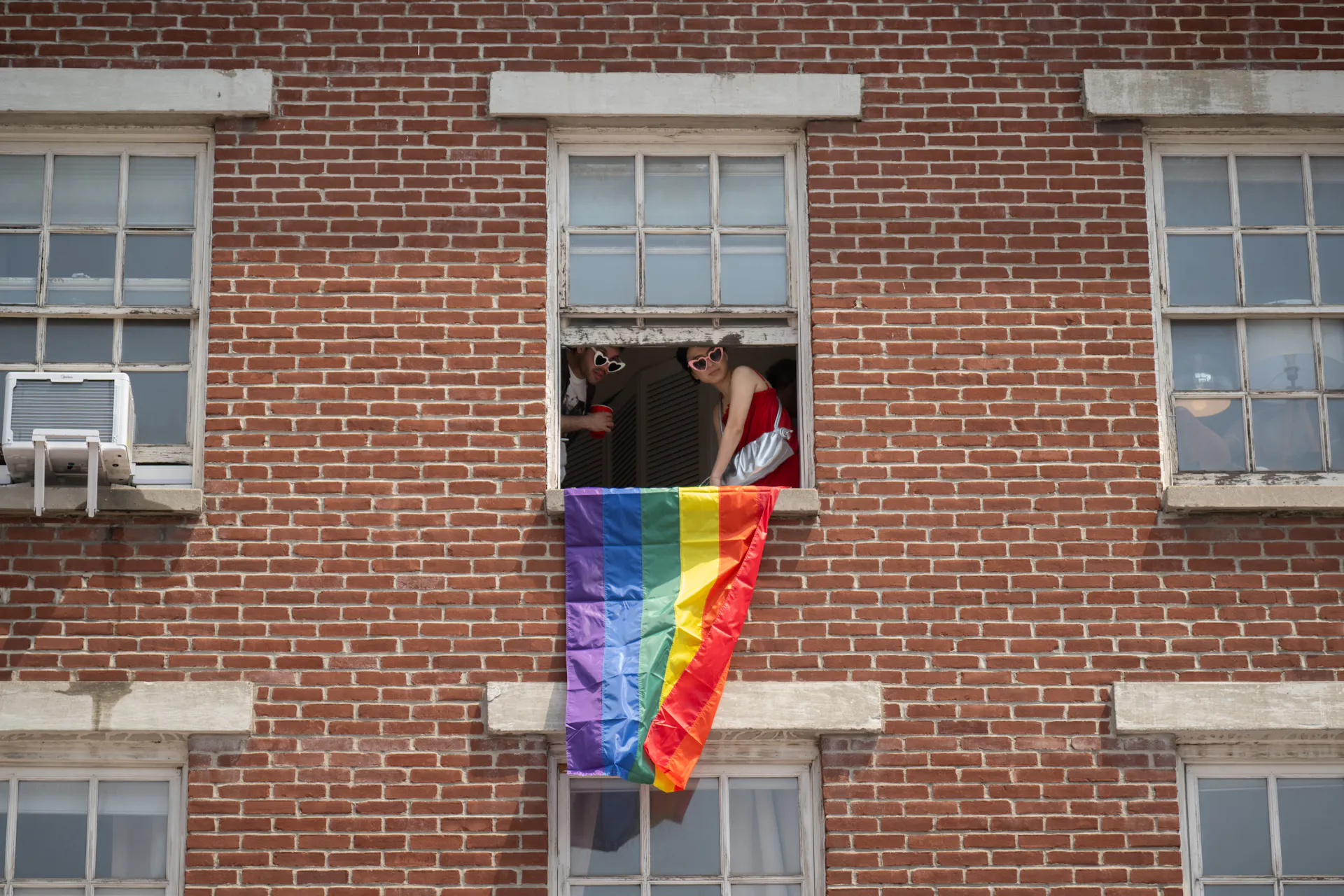 Two people wearing heart-shaped sunglasses lean out a window of a red brick building, a rainbow flag hanging from the sill.