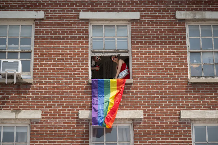 Two people wearing heart-shaped sunglasses lean out a window of a red brick building, a rainbow flag hanging from the sill.