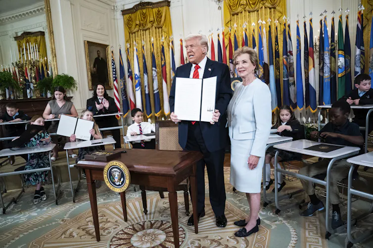 President Trump poses with a woman in a dressy suit; he is holding two big pieces of paper and there are flags and children in the background seated at little desks.
