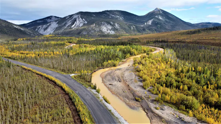 un río beige cerca de una carretera en una zona forestal con una cadena montañosa rocosa en la distancia