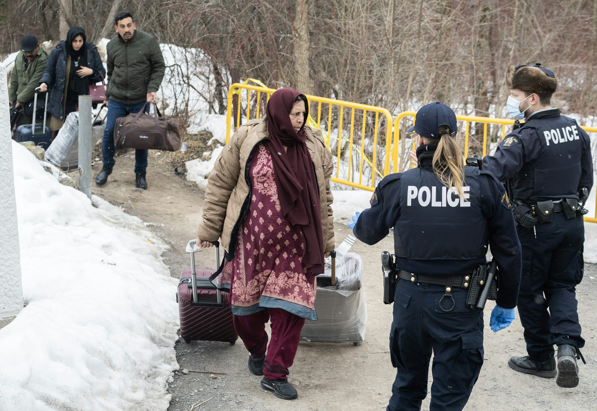 Una mujer cruza la frontera con bolsos y una maleta y es recibida por dos agentes de la RCMP con mascarillas.