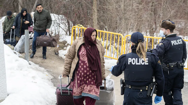 Una mujer cruza la frontera con bolsos y una maleta y es recibida por dos agentes de la RCMP con mascarillas.