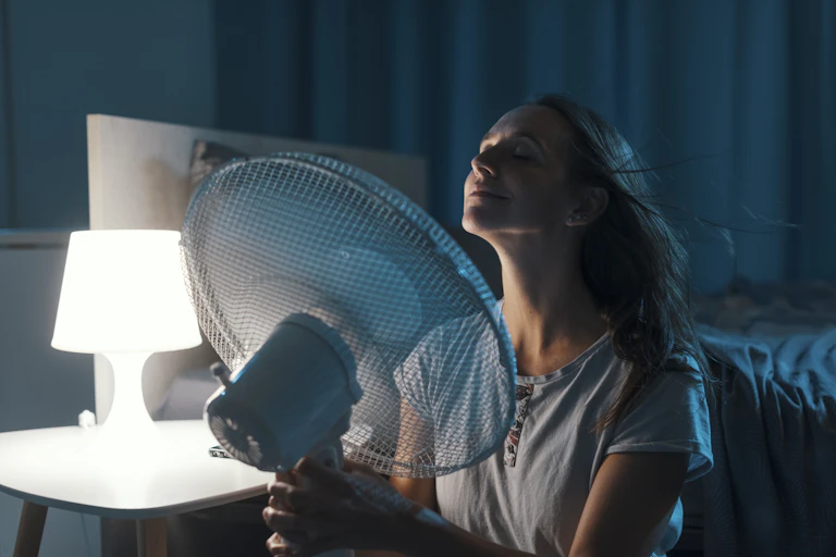 Una mujer en la habitación de noche sujeta un ventilador delante de su cara junto a la mesita de noche con una lámpara encendida