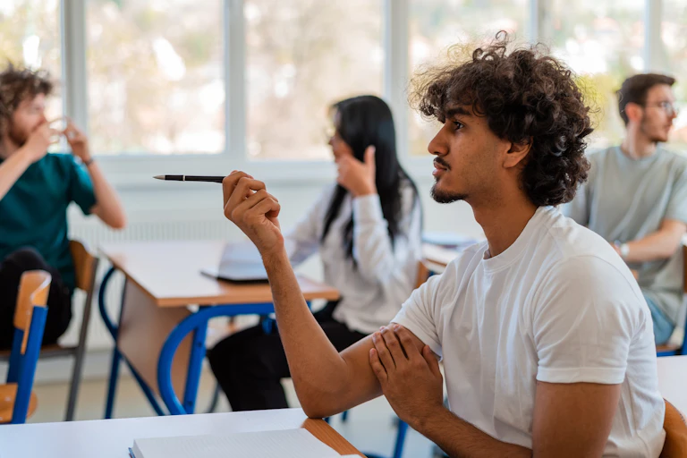 In a classroom, university students are chatting amongst themselves and one of them is pointing with a pen