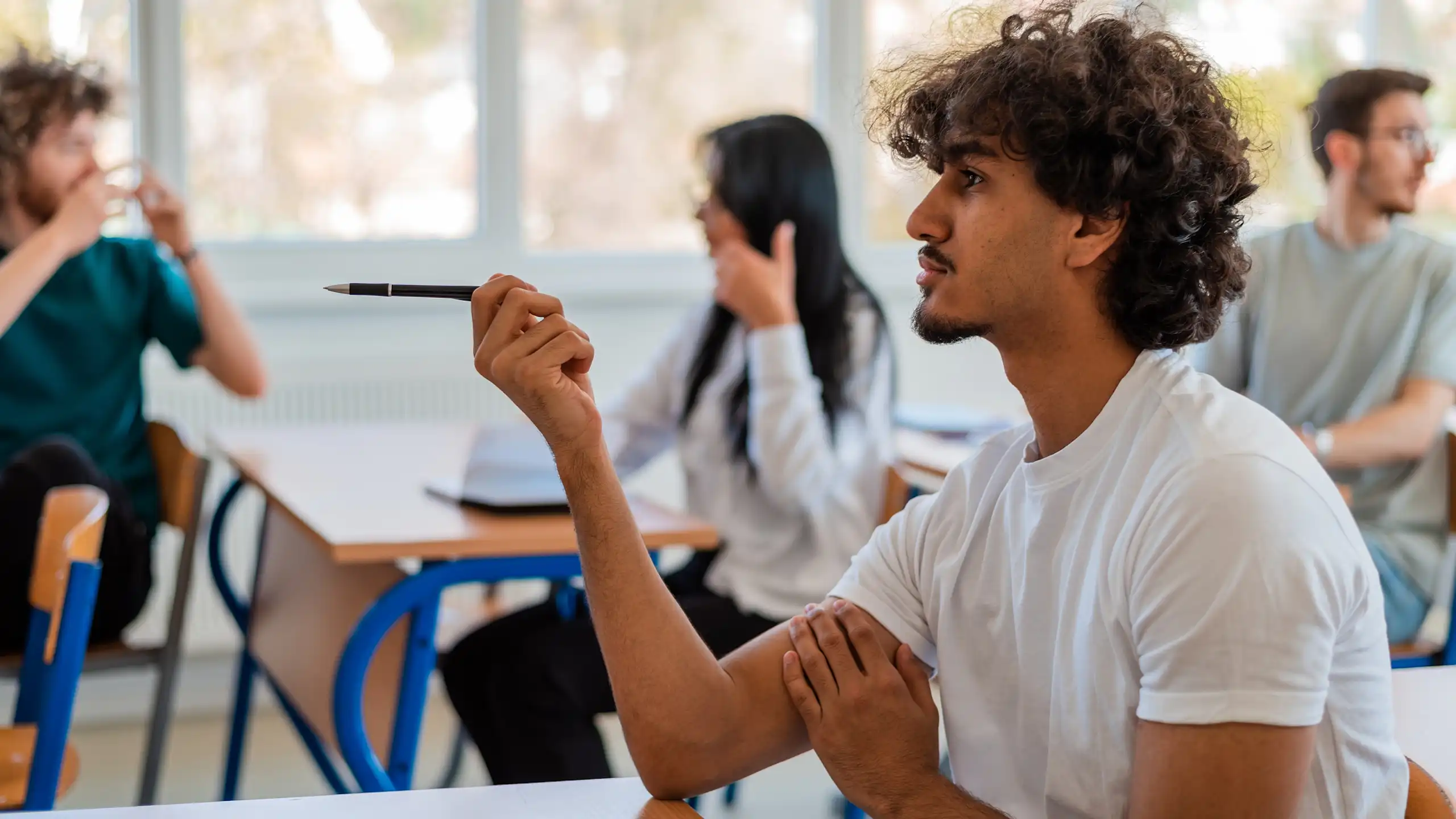 In a classroom, university students are chatting amongst themselves and one of them is pointing with a pen