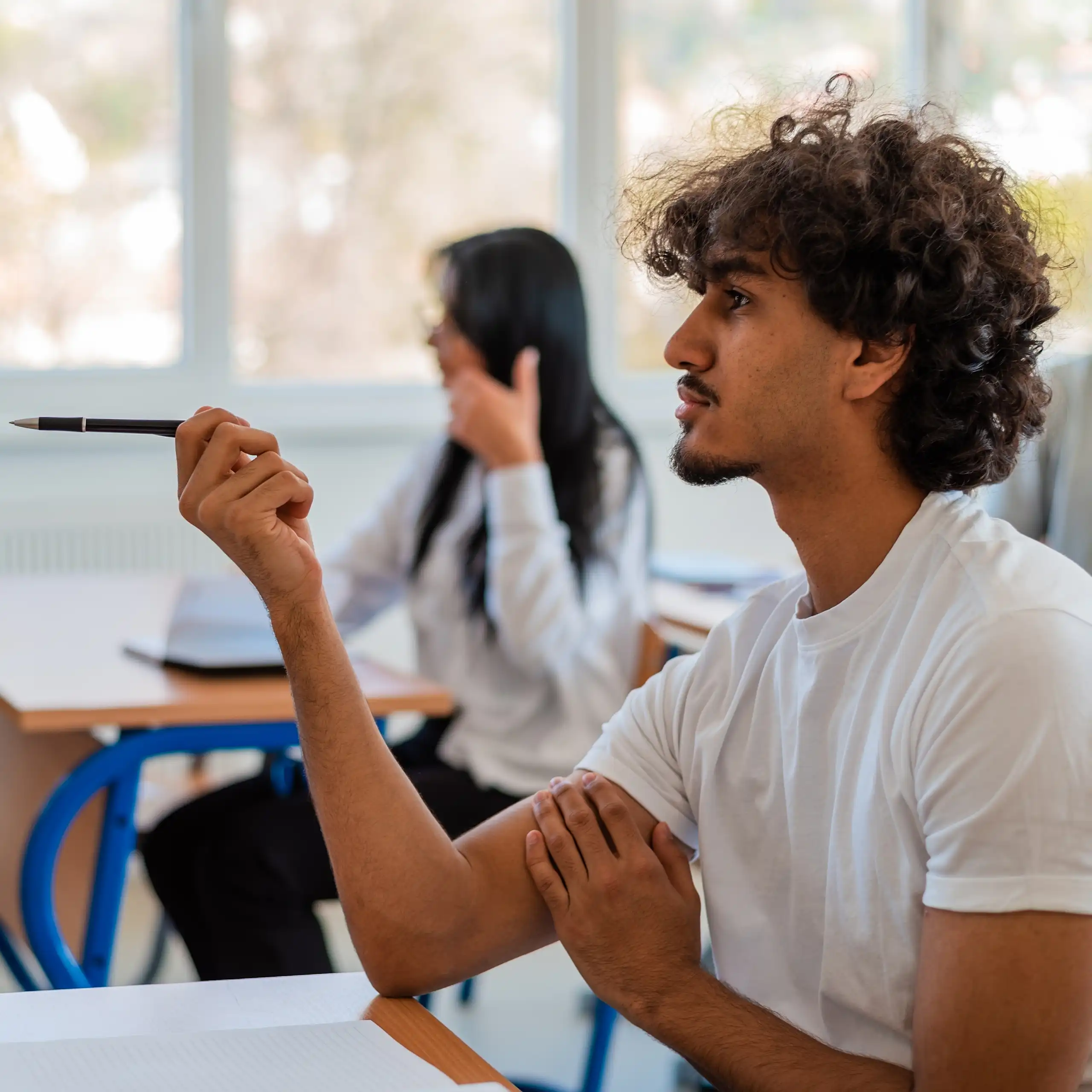 In a classroom, university students are chatting amongst themselves and one of them is pointing with a pen