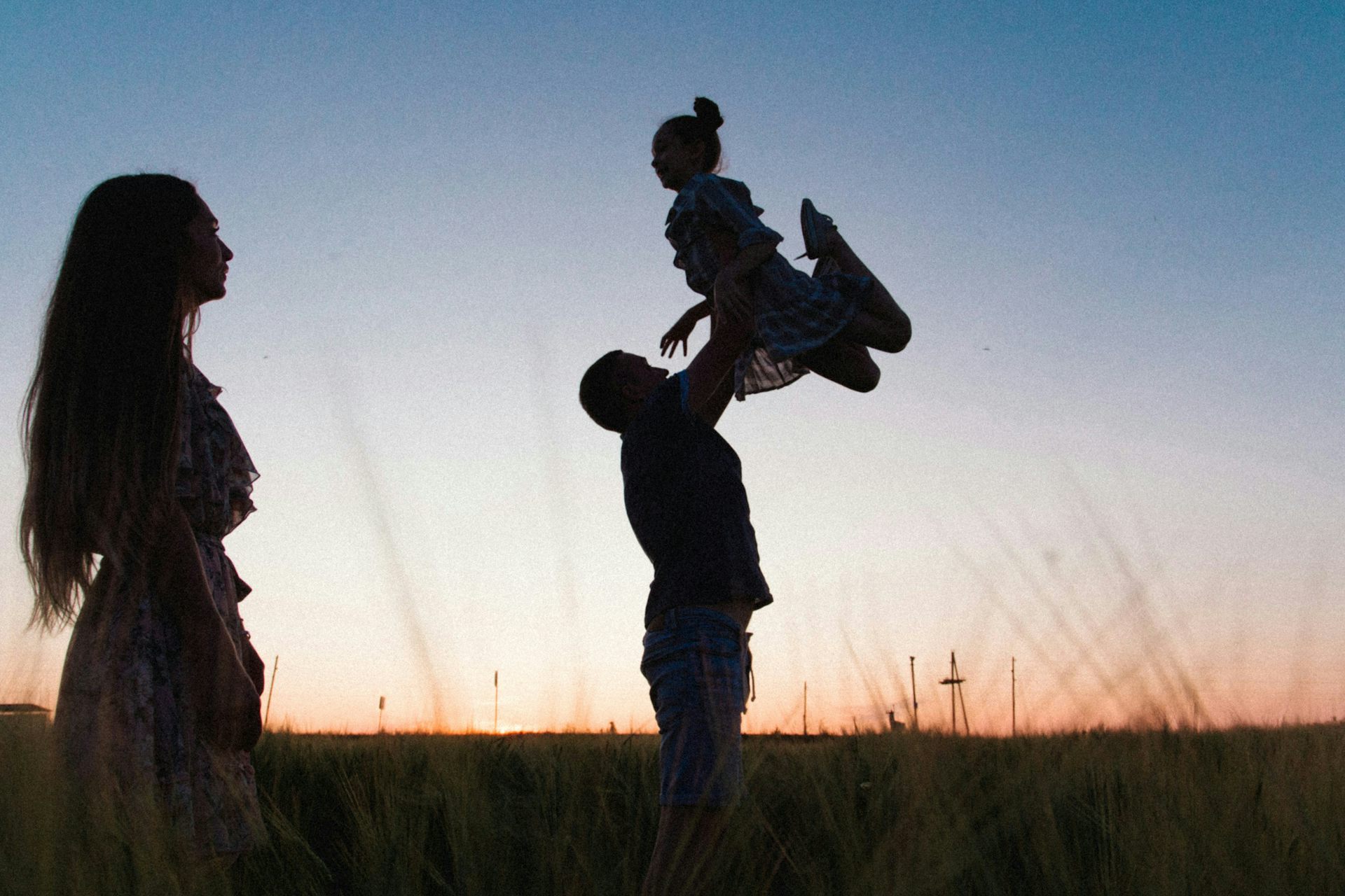 a man tosses a girl in the air at sunset in silhouette as a woman looks on