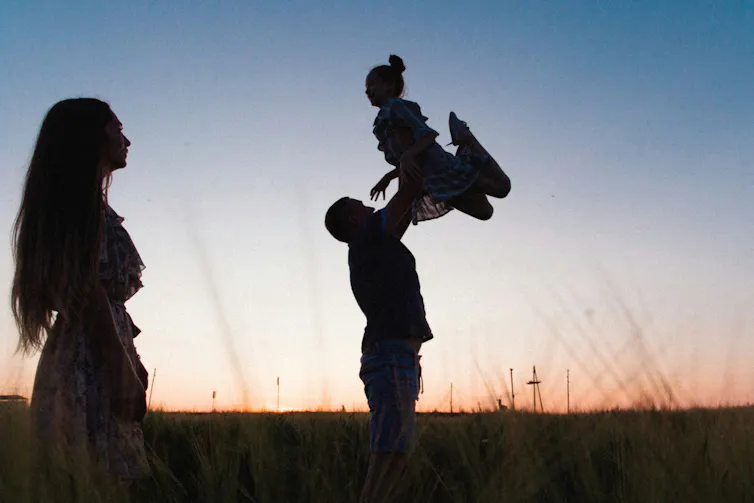 a man tosses a girl in the air at sunset in silhouette as a woman looks on