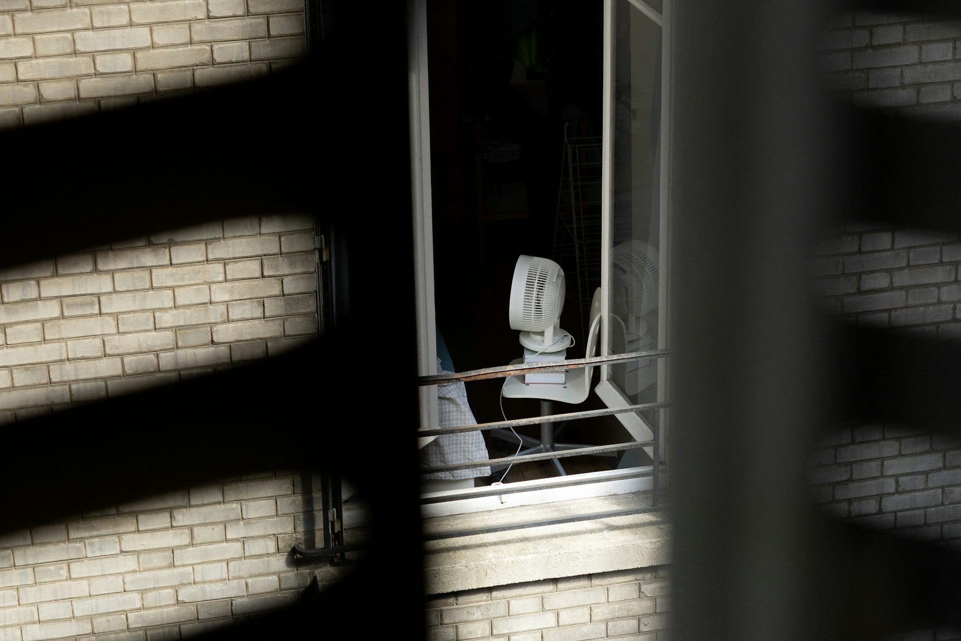 Vue d'une fenêtre sur cour sur un appartement où un ventilateur est posé sur une chaise, tournée vers un lit.