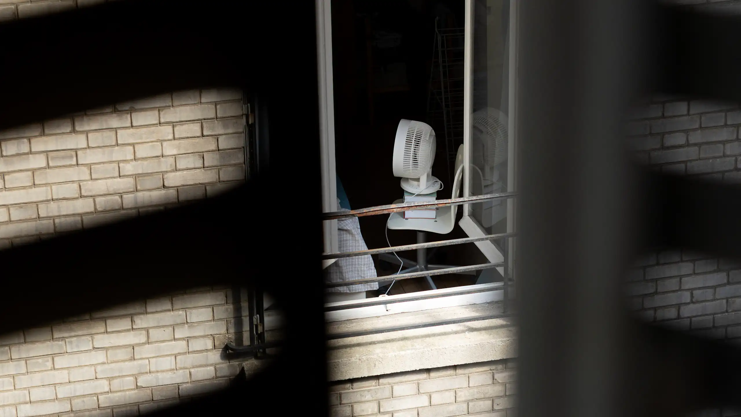 Vue d'une fenêtre sur cour sur un appartement où un ventilateur est posé sur une chaise, tournée vers un lit.