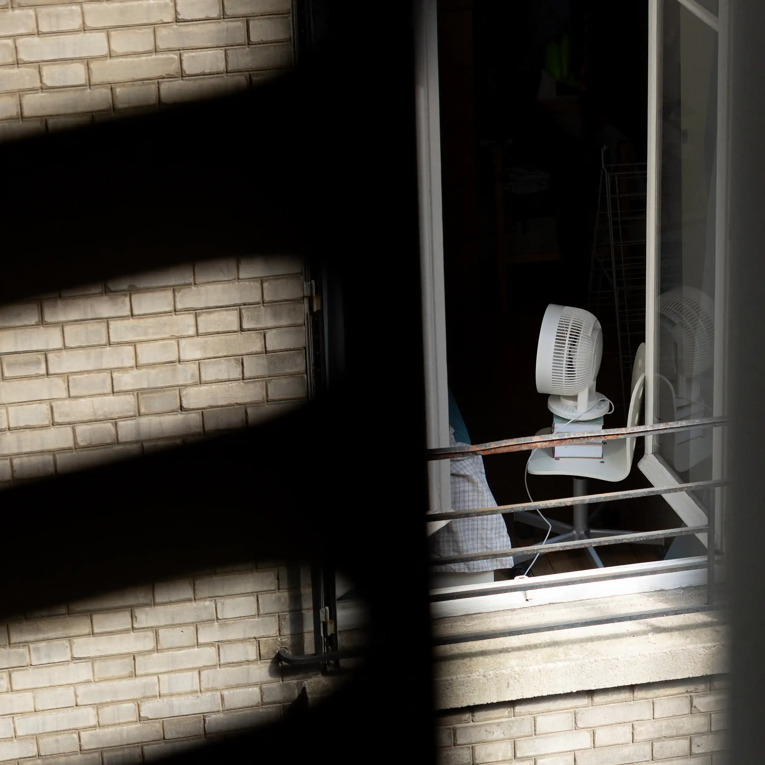 Vue d'une fenêtre sur cour sur un appartement où un ventilateur est posé sur une chaise, tournée vers un lit.