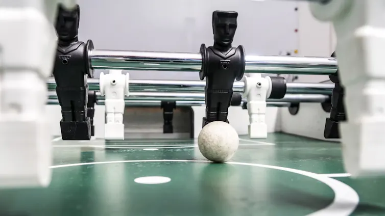 A foosball set with toy figures on metal rods used to propel a small football, a row of black figures on strike, white figures foregrounded.