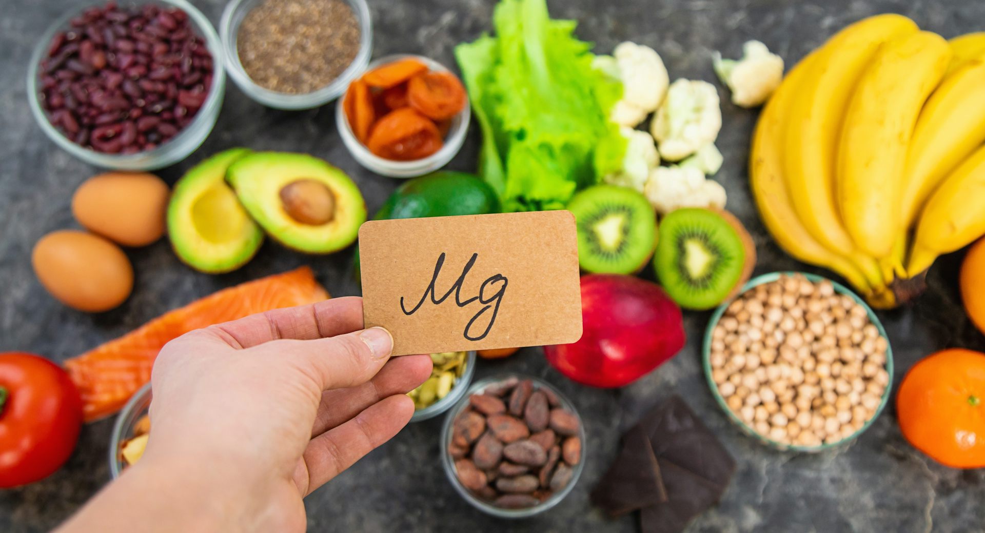 Uma mão segura uma folha de papel na qual está escrito “Mg”, em frente a frutas e legumes.