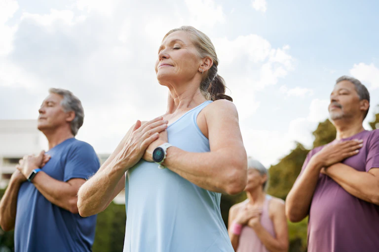 Seniors meditating outdoors in a park with hands on their heart and eyes closed.