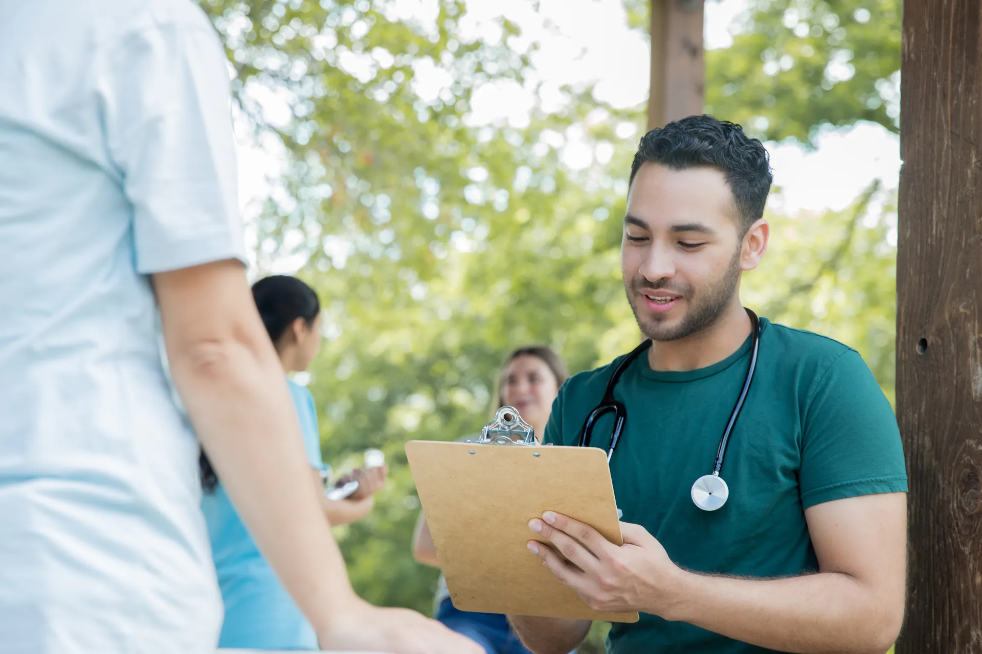 A male doctor or nurse with a stethoscope around his neck writes on a clipboard while talking to someone at a community health event