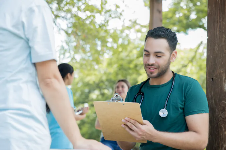 Un médico o enfermero con un estetoscopio alrededor del cuello escribe en un portapapeles mientras habla con alguien en un evento de salud comunitario